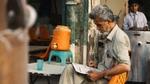 Older man sits outdoors reading a newspaper beside a water cooler and plastic cup.
