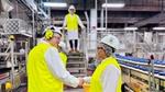 Two factory workers in yellow high-vis jackets and hard hats shake hands next to a Unilever factory production line.