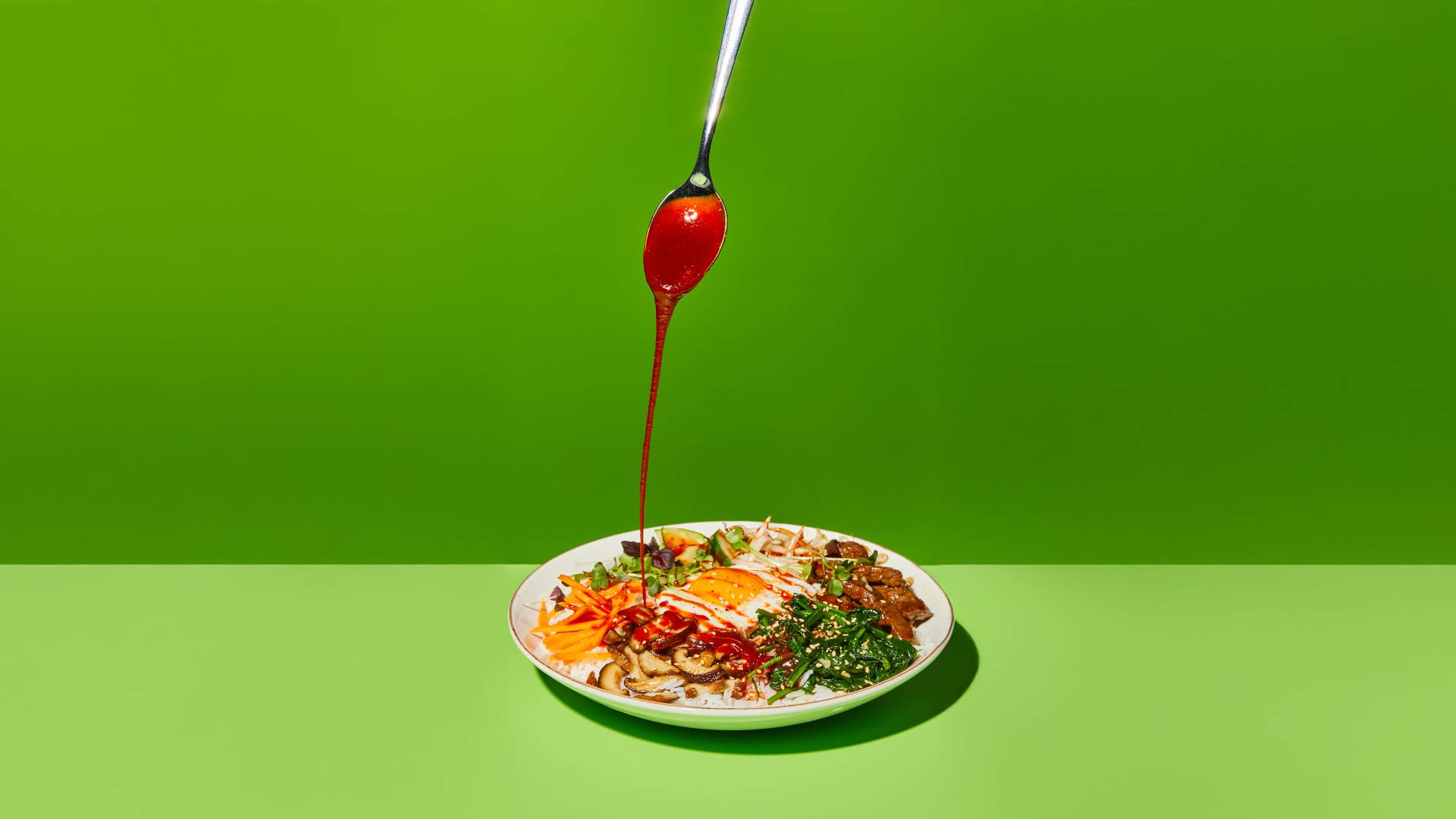 A red sauce is drizzled from a teaspoon over a colourful dish of assorted ingredients topped with a fried egg.