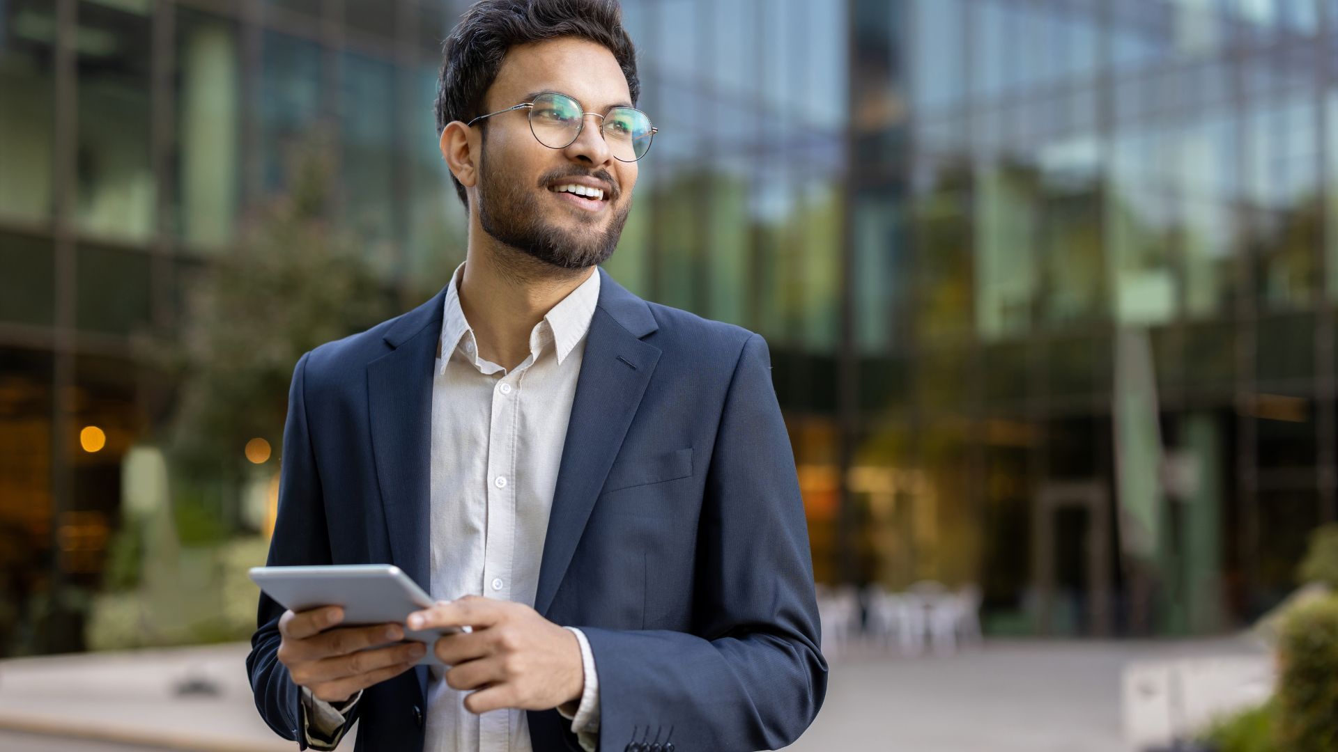Smiling man in a suit holding a tablet outside a glass building.