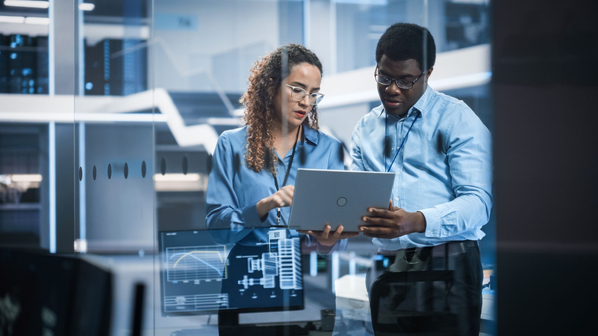 Two professionals reviewing data on a laptop in a modern, high-tech office with monitors displaying graphs in the foreground. They are both wearing glasses and dressed in blue shirts.