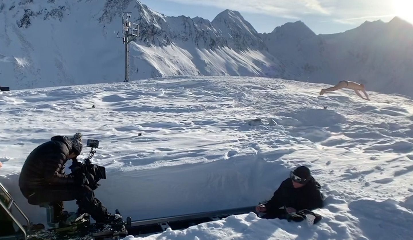 On a sunny snow field surrounded by mountains  a photo crew ski wear is photographing a man doing press-ups in boxer shorts