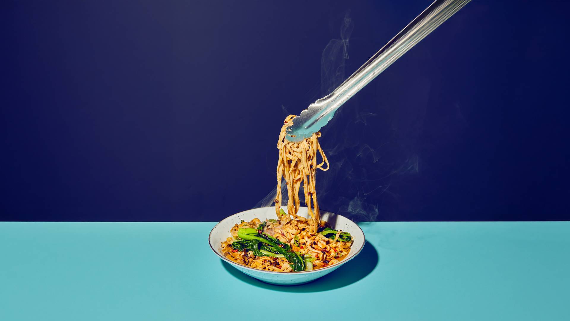 An image of Asian-inspired noodles being lifted from a white bowl  with tongs, shot against a split background of dark and light blue.
