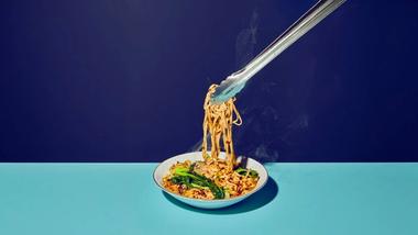 An image of Asian-inspired noodles being lifted from a white bowl with tongs, shot against a split background of dark and light blue.
