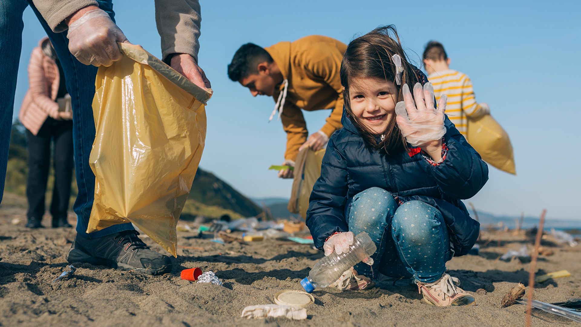 A young girl holding a rubbish bag, waving as she collects rubbish off a beach