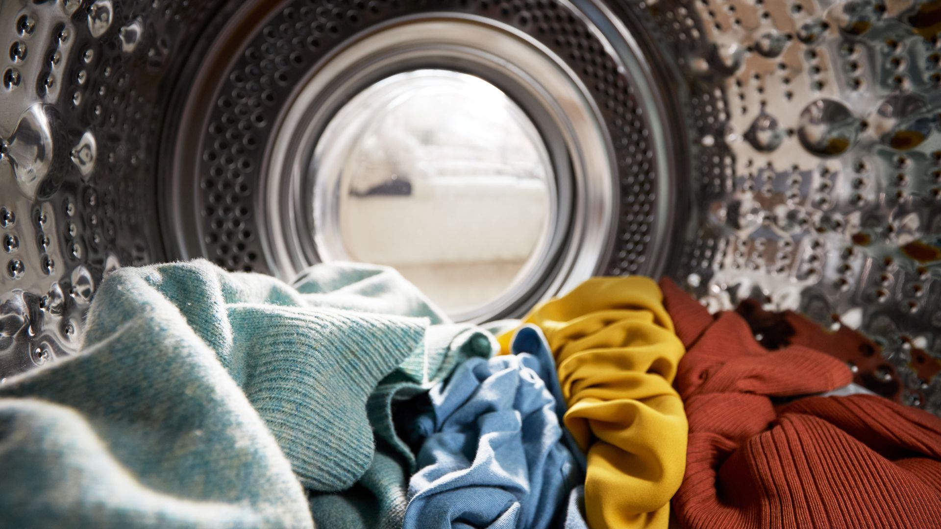 Close-up view from inside a washing machine drum, showing laundry in shades of green, blue, yellow and red ready to be washed.
