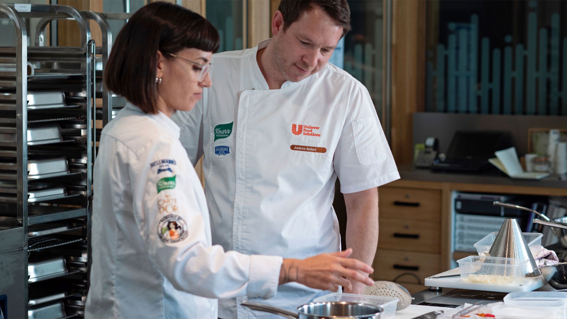 A male and female chef wearing their uniforms chatting in a kitchen