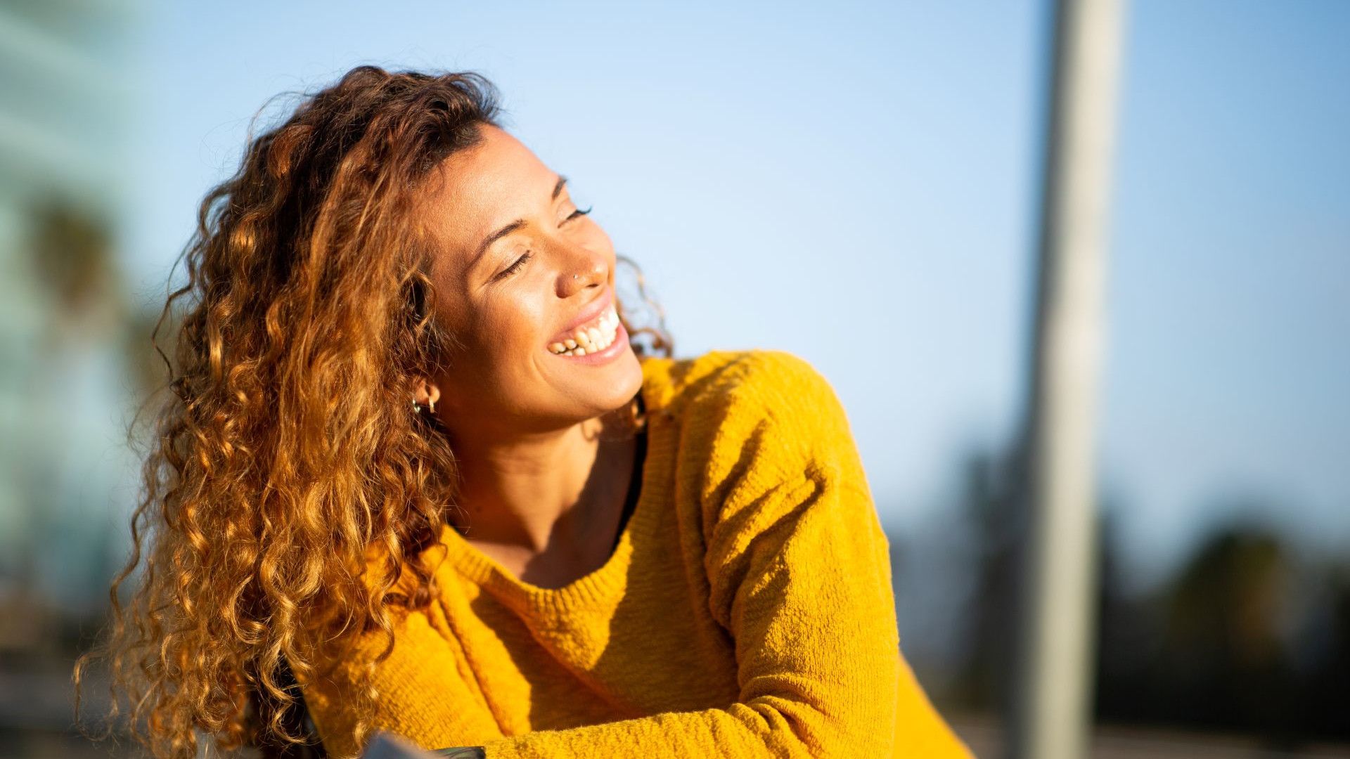 Woman in orange jumper, standing in a flowery meadow with bright blue sky and a dry stone wall in the background.
