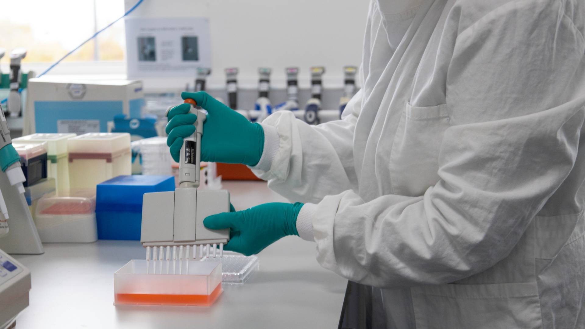 A scientist at work in a Unilever R&D lab. Their face is not visible. They are wearing a white lab coat and green gloves, holding a device.