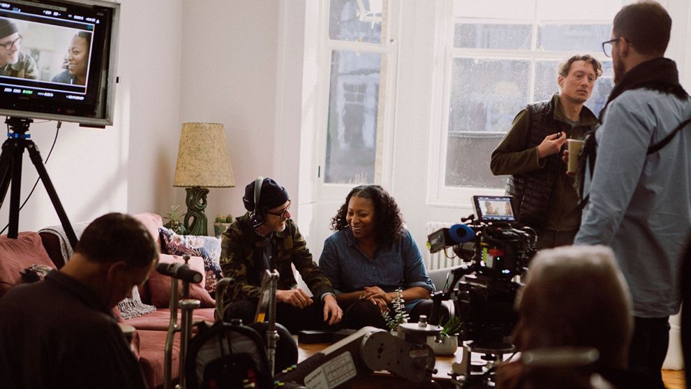 A film crew is taking a break in a light-filled living room with camera equipment on a low table and a screen on a tripod.