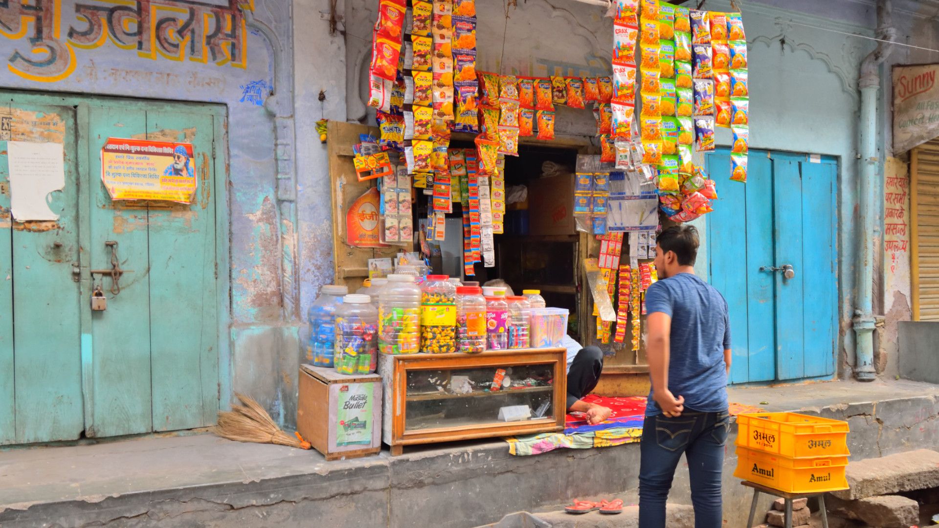 A man shops at a small store in rural India. Colourful goods are displayed on a table and hanging from the wall.