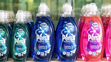 Persil Wonder Wash plastic laundry detergent bottles lined up in a row. There are three colours of detergent – green, blue and pink. The packaging labels explain that the product is designed for short cycles.